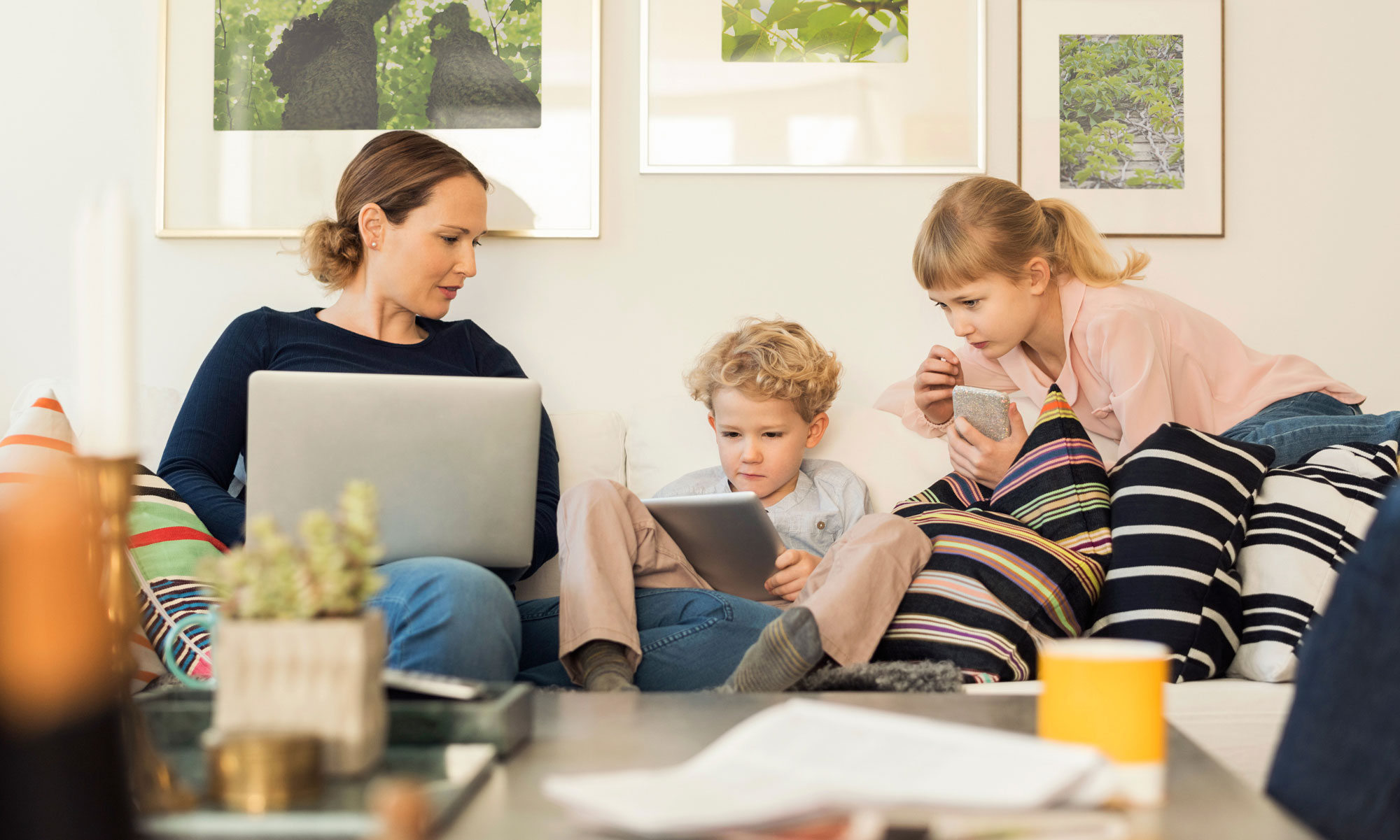 Mamma och två barn sitter i soffa och tittar på olika skärmar